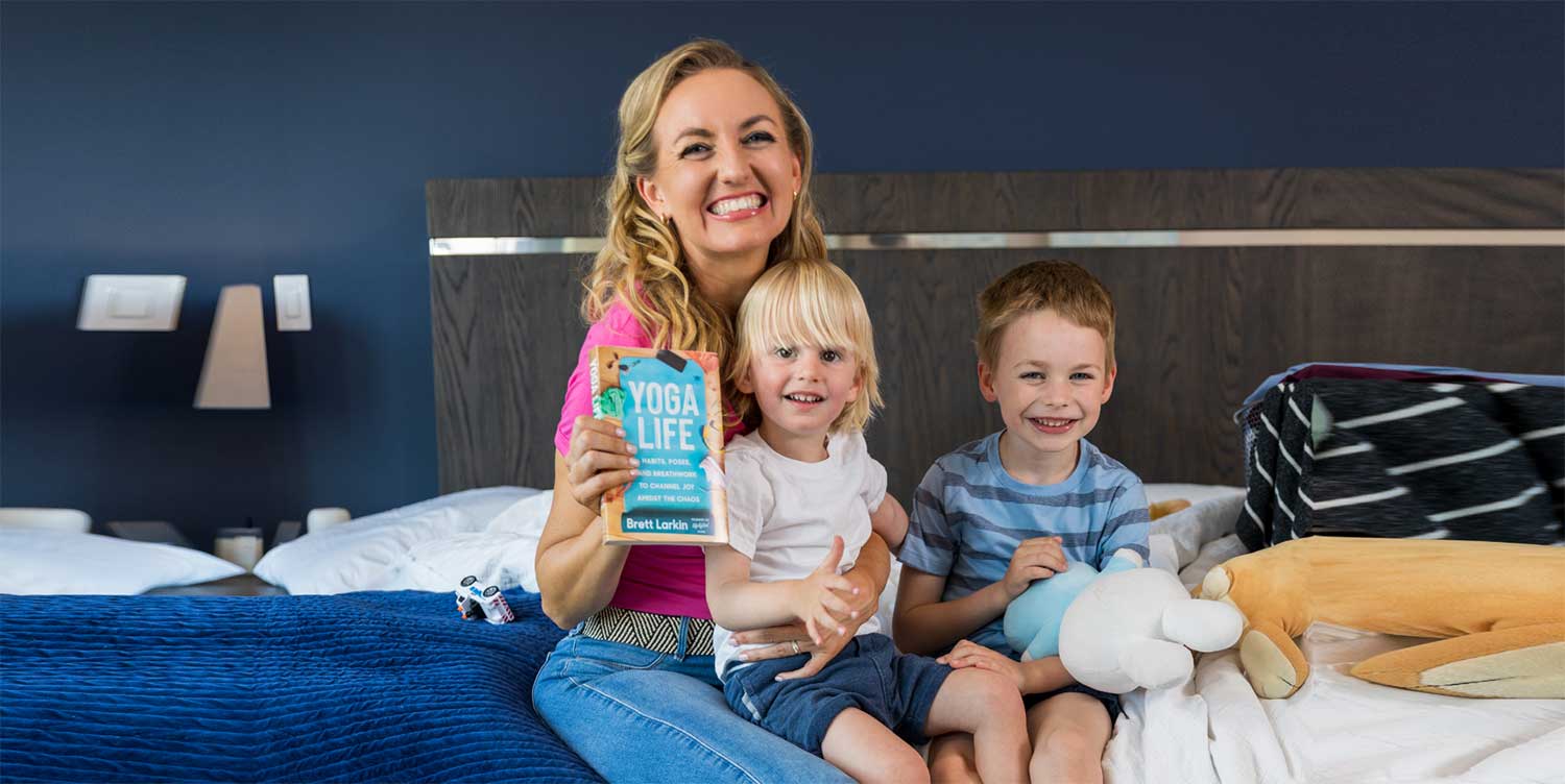 Brett Larkin sitting on her bed with her two young children, surrounded by the chaos of laundry and toys, and holding her book, Yoga Life.