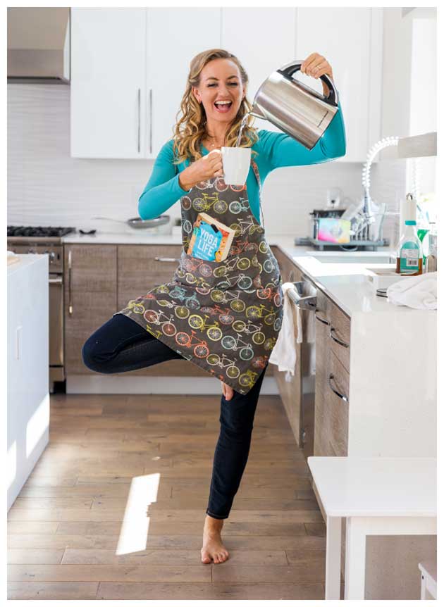 Brett Larkin practicing tree pose in the kitchen while pouring herself a cup of tea and carrying her book, Yoga Life, in the pocket of her apron.