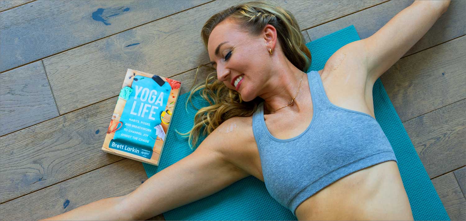 Brett Larkin laying with arms stretched out on a yoga mat and facing toward her book, Yoga Life.