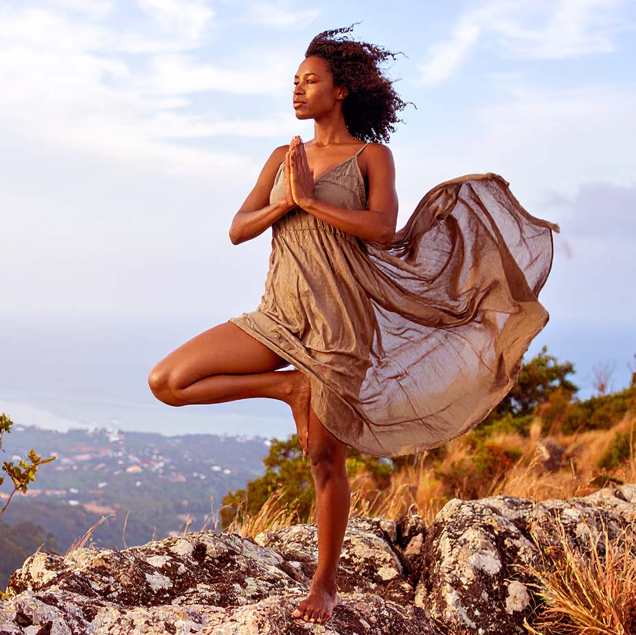 Woman practicing tree pose outdoors with hands in prayer position, representing meditation, balance, and Kundalini Yoga practice.