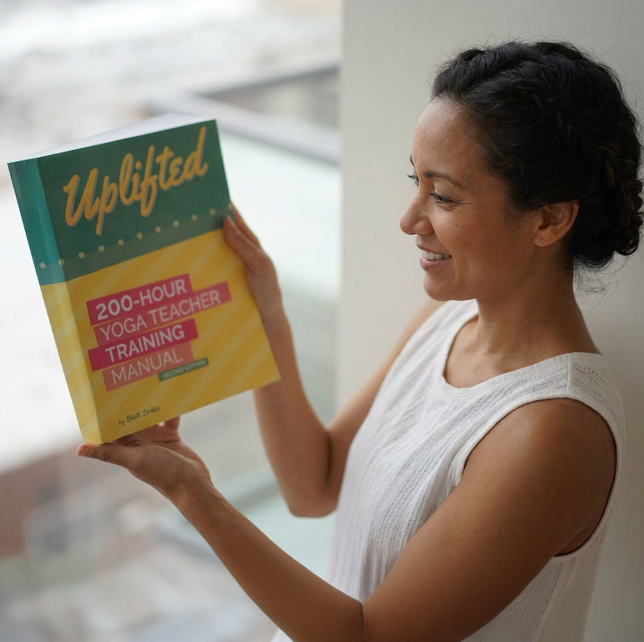 Student holding the Uplifted 200-Hour Yoga Teacher Training Manual during an online yoga teacher certification program.